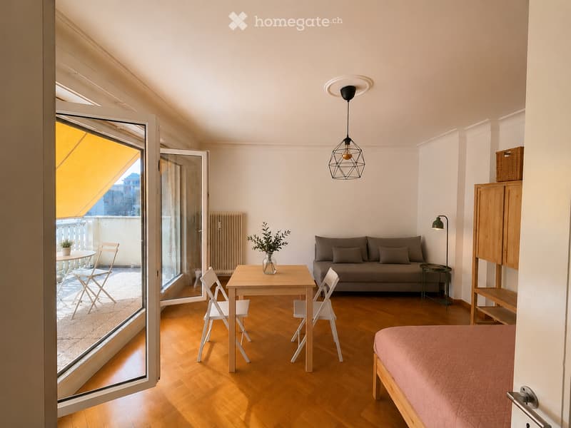 Bright living space with wooden flooring, a sofa, a small dining table with chairs, a bed, and a balcony with outdoor furniture.
