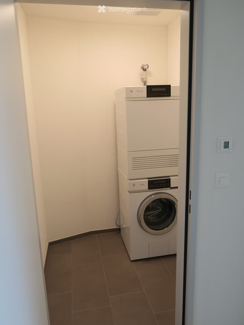 A small, well-lit room with a stacked washing machine and dryer unit against one wall, tiled floor, and white walls.
