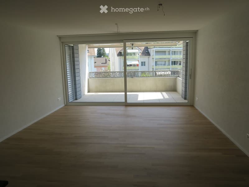 Empty room with wooden flooring and large sliding glass doors leading to a balcony with a view of neighboring buildings.