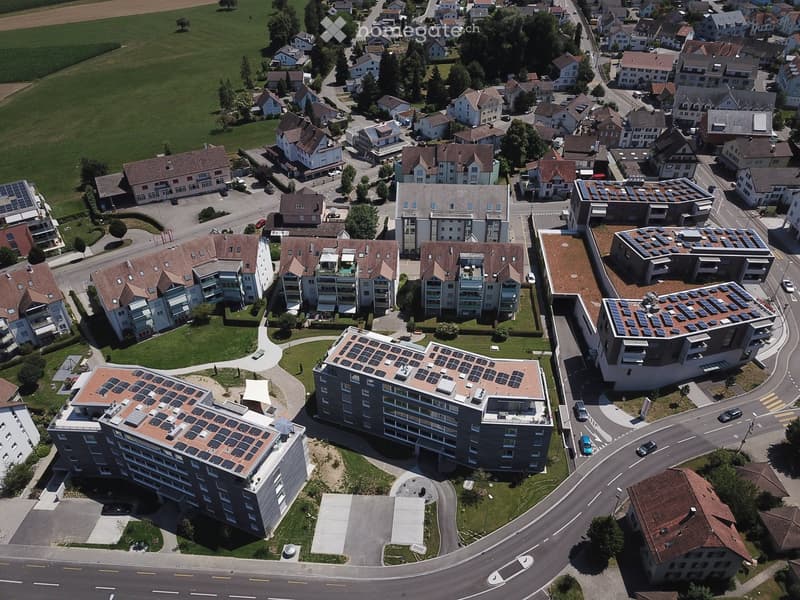 Aerial view of a residential area with multiple buildings, including apartment complexes with solar panels on the roofs, surrounded by green spaces and roads.