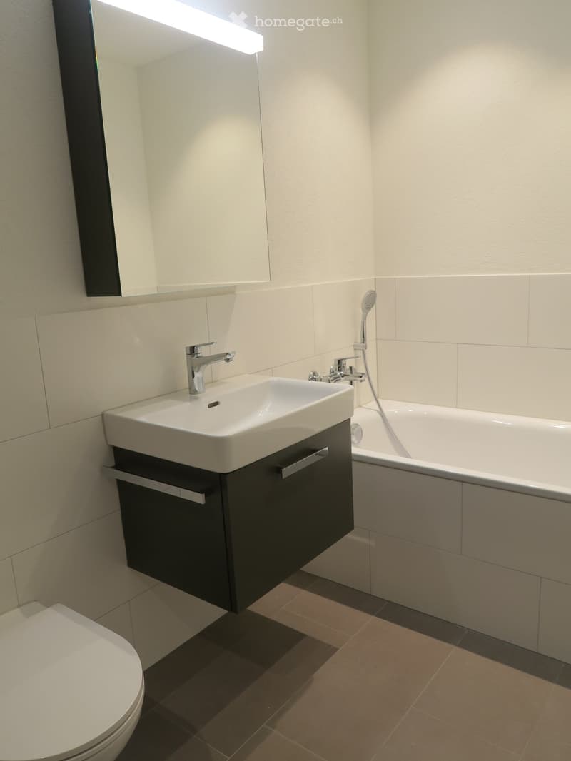 Modern bathroom with a white sink, chrome faucet, black cabinet, bathtub, and a toilet. The walls are tiled in light beige, and the floor is tiled in brown.