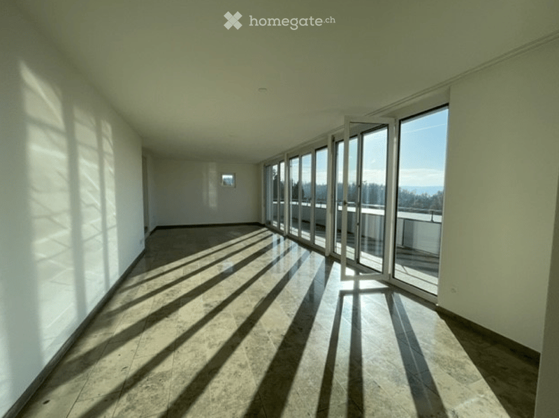 Bright, empty room with large floor-to-ceiling windows and sliding glass doors leading to a balcony. Light-colored tiled floor and white walls.