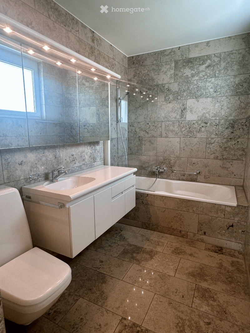 Modern bathroom with a white toilet, sink, and bathtub. Features a large mirror with integrated lighting, granite tiles on walls and floor, and a glass-enclosed shower area.