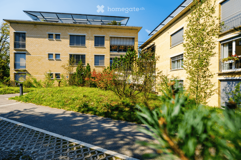 Two-story residential building with yellow brick facade, multiple windows, and a rooftop terrace. The building is surrounded by greenery and a paved pathway.