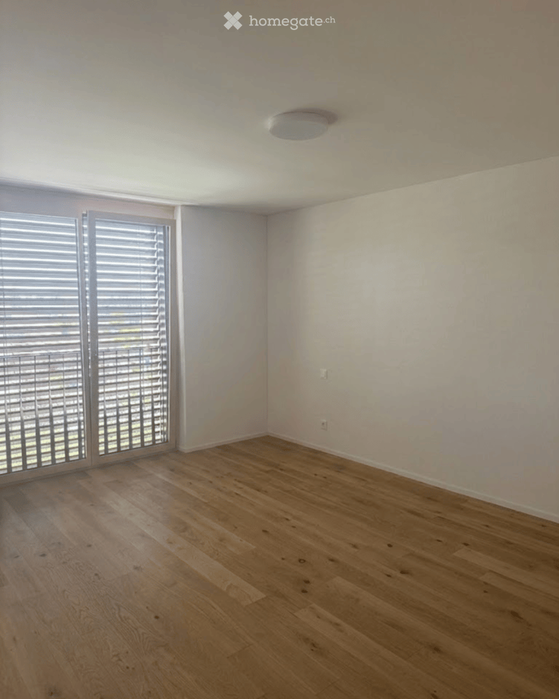 Empty room with wooden flooring, white walls, large window with blinds, and a ceiling light fixture.