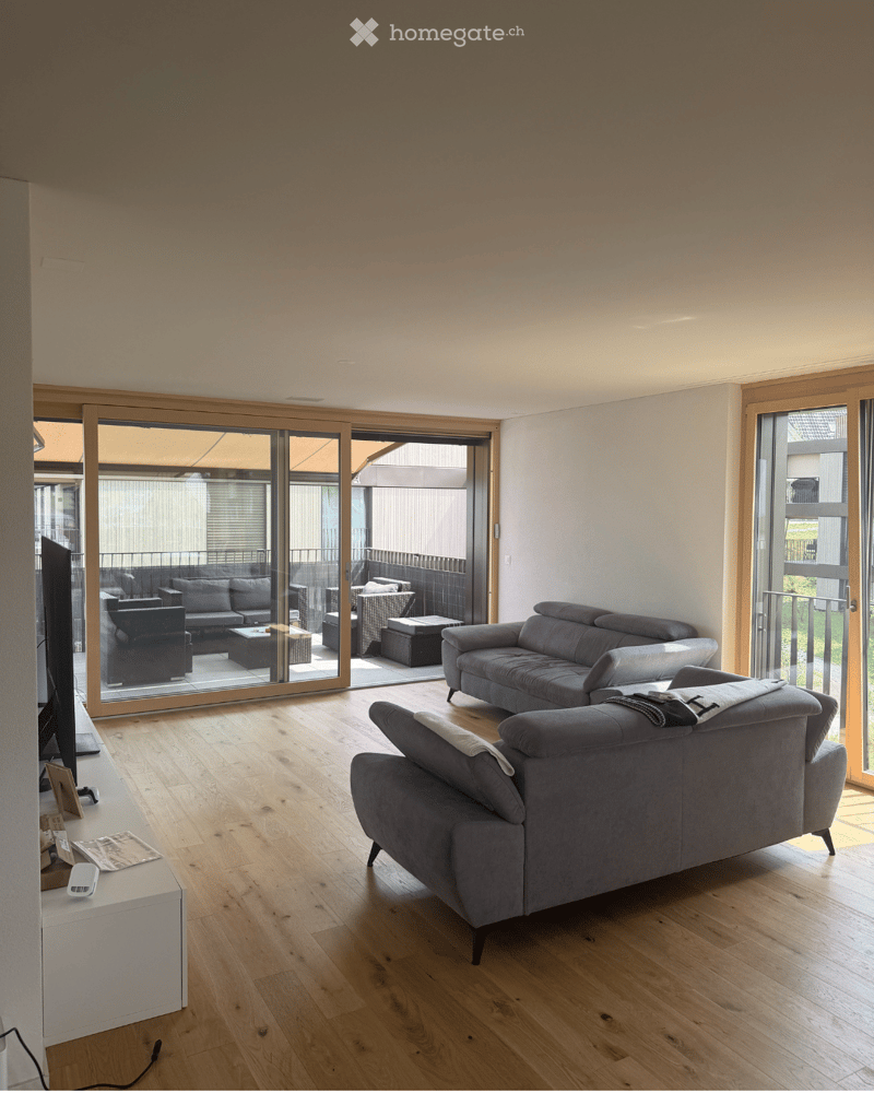Modern living room with light wooden flooring, large sliding glass doors leading to a balcony, grey sectional sofa, coffee table, armchairs, ottoman, and a TV screen on a stand.