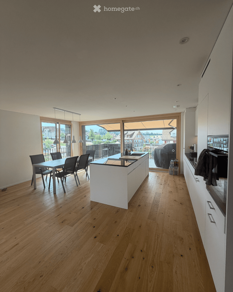Modern kitchen with light wooden flooring, white cabinetry, a central island, and a dining area with a table and chairs. Large windows provide natural light and a view of the outdoor area.