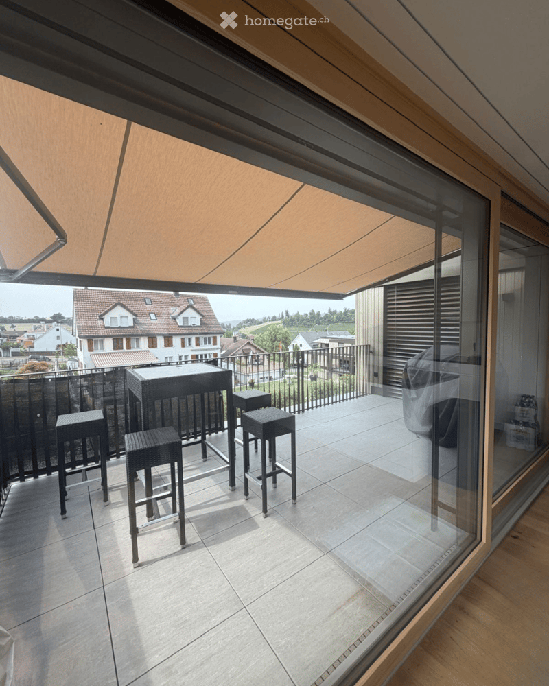 Balcony with tiled floor, black metal railing, and a table with four stools, covered by a retractable awning.