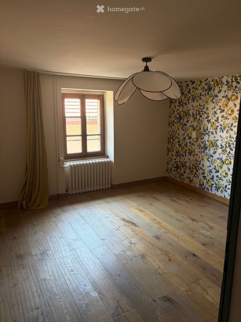 Empty room with wooden flooring, floral wallpaper, a window with a curtain, a radiator, and a modern pendant light fixture.