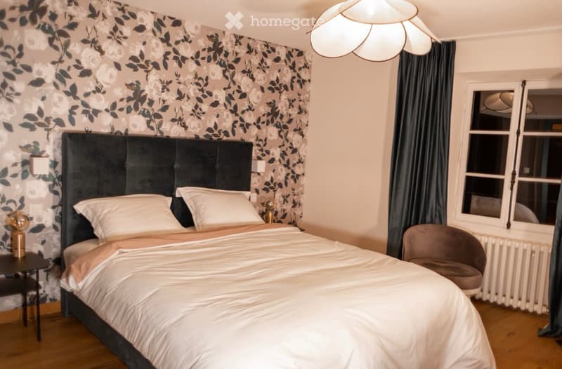 Cozy bedroom with a large upholstered headboard, white bedding, a nightstand with a lamp, and a small armchair near the window.