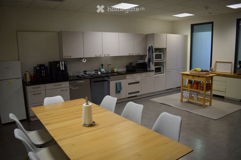Modern kitchen with light grey cabinets, stainless steel appliances, a wooden dining table with white chairs, and a small cart with drinks and snacks.