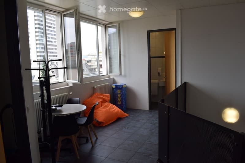 A small, furnished room with a round table, chairs, a radiator, a screen, and a clothing rack near the window. An orange bean bag is placed on the floor, and a blue recycling bin is visible.