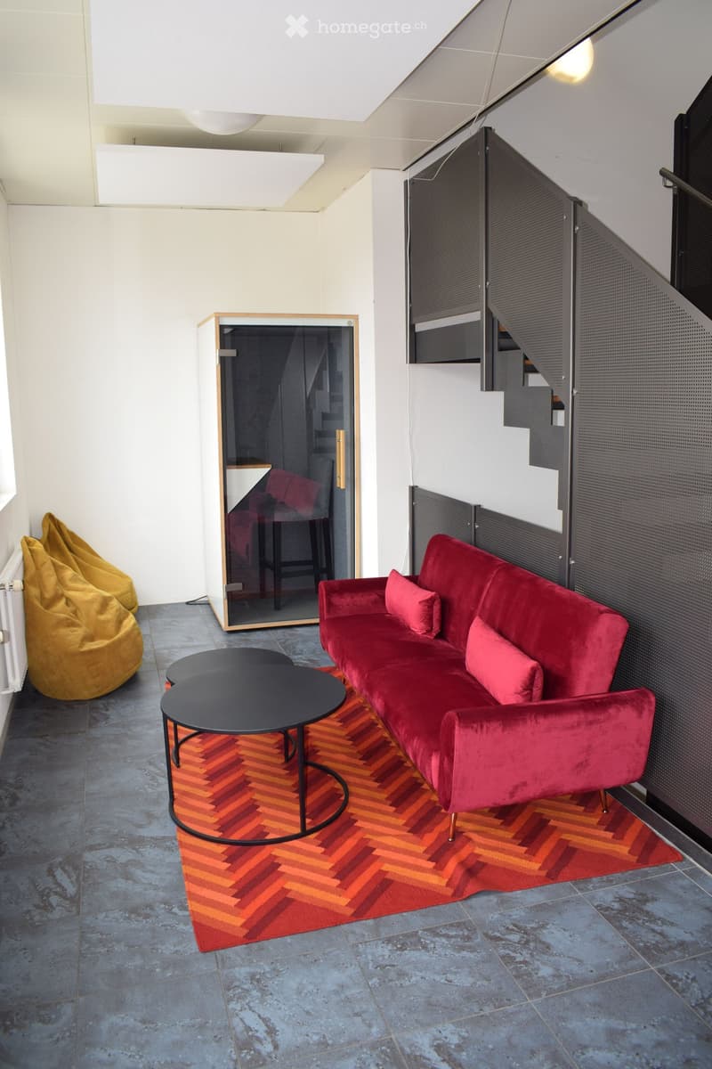 Modern living room with a red velvet sofa, black coffee table, and bean bag chairs. Features a geometric patterned rug and a staircase leading upwards.