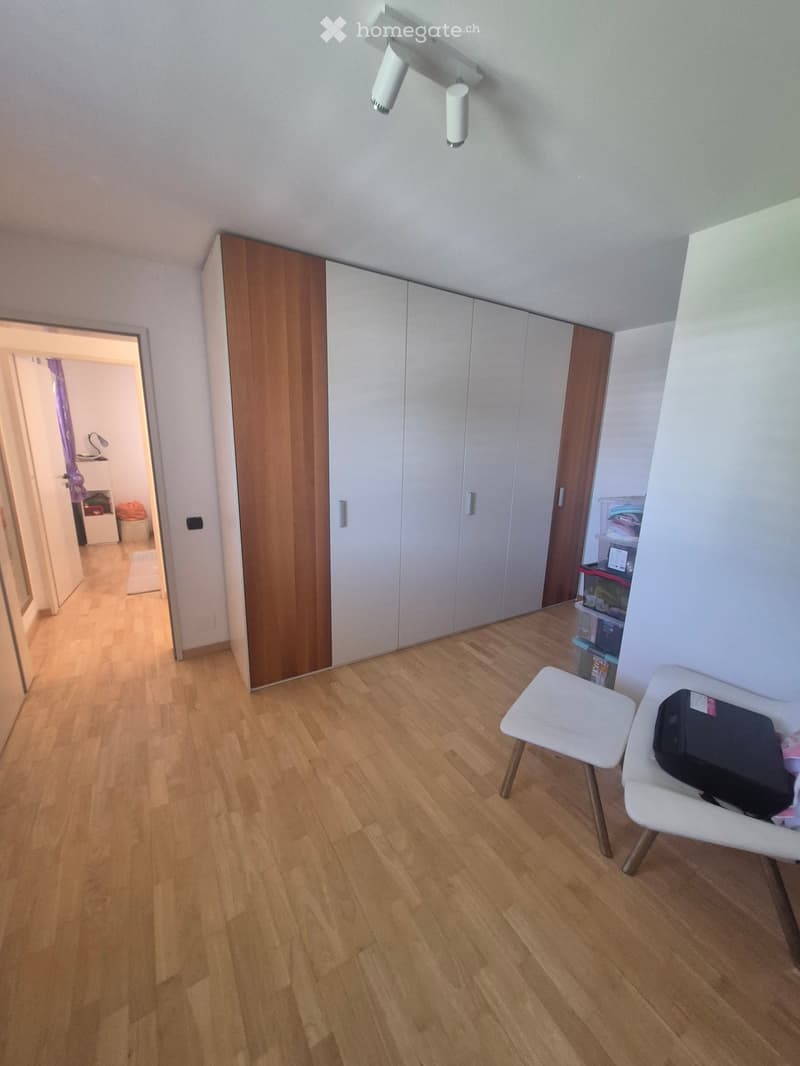 A small, well-lit hallway with wooden flooring, a white wardrobe, a small white stool, and a white chair with a black bag on it.