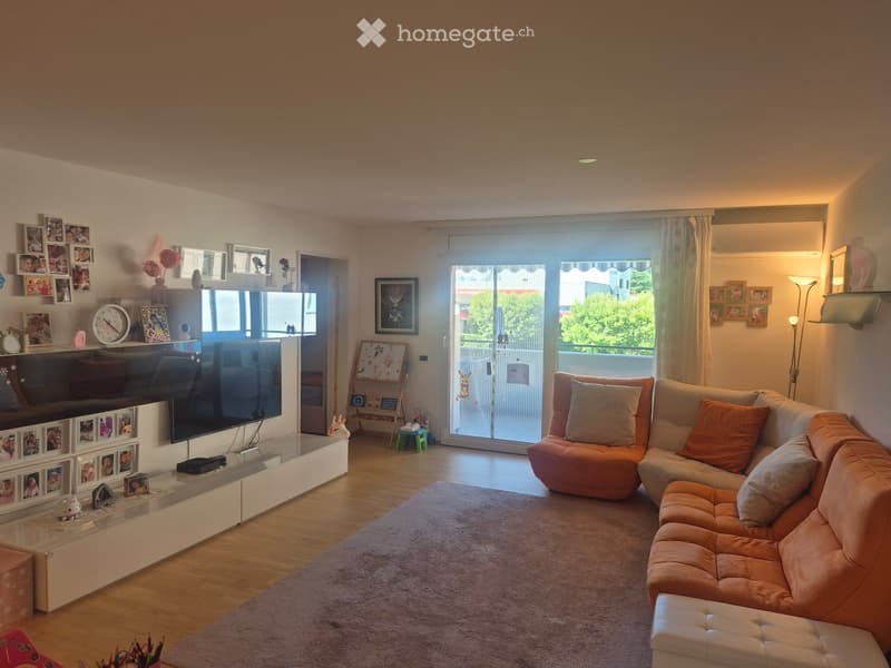 Modern living room with a large sectional sofa, coffee table, TV, floor lamp, and decorative picture frames. Large windows offer a view of greenery outside.