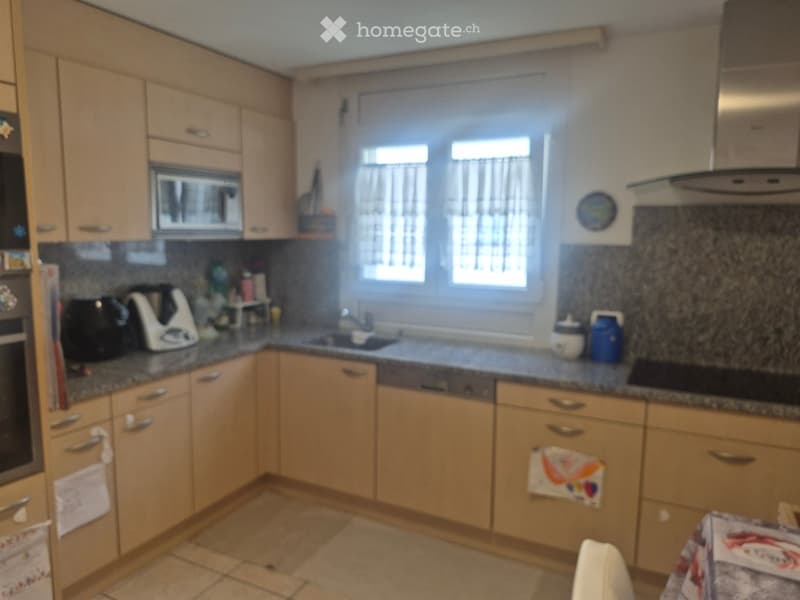 Well-lit kitchen with light wooden cabinets, granite countertops, and various appliances including a microwave, oven, fridge, and blender. There is a sink with a faucet, a dishwasher, and a stove. The floor is tiled, and there are decorative towels hangin