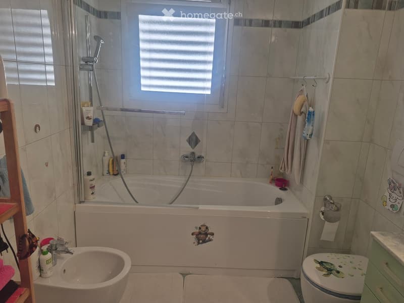 Bathroom with a bathtub, shower, sink, and toilet. Features tiled walls, a window with blinds, and various toiletries on shelves.
