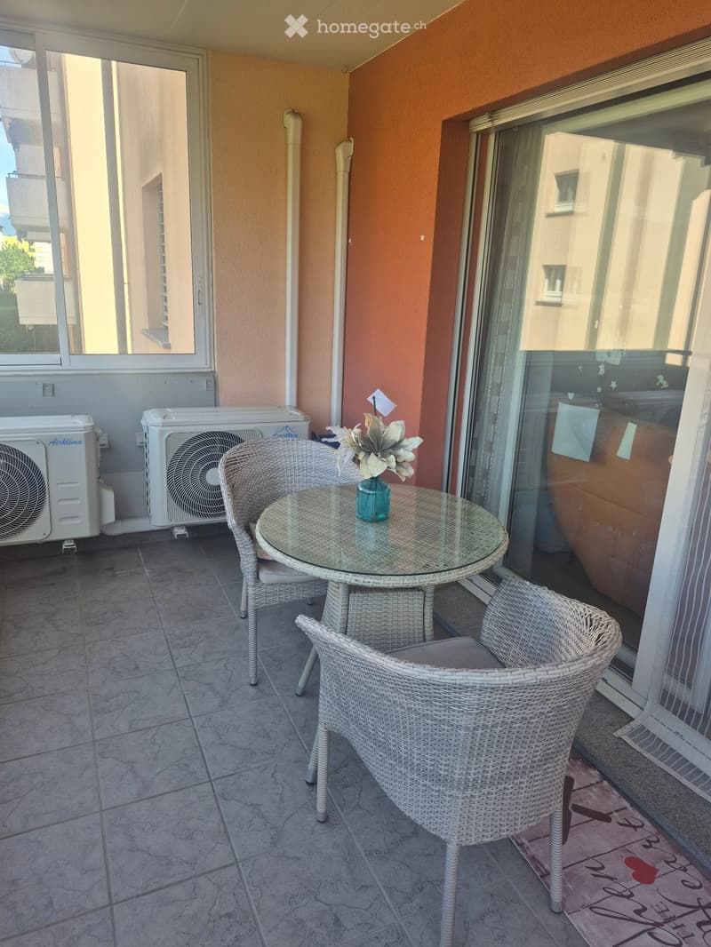 Balcony with wicker chairs, a glass table, and a vase with flowers. Air conditioning units are visible on the wall.