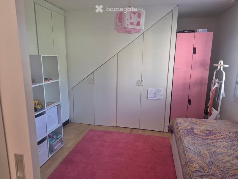 A well-lit bedroom with a bed, pink wardrobe, white shelving unit, and a red rug on the floor.