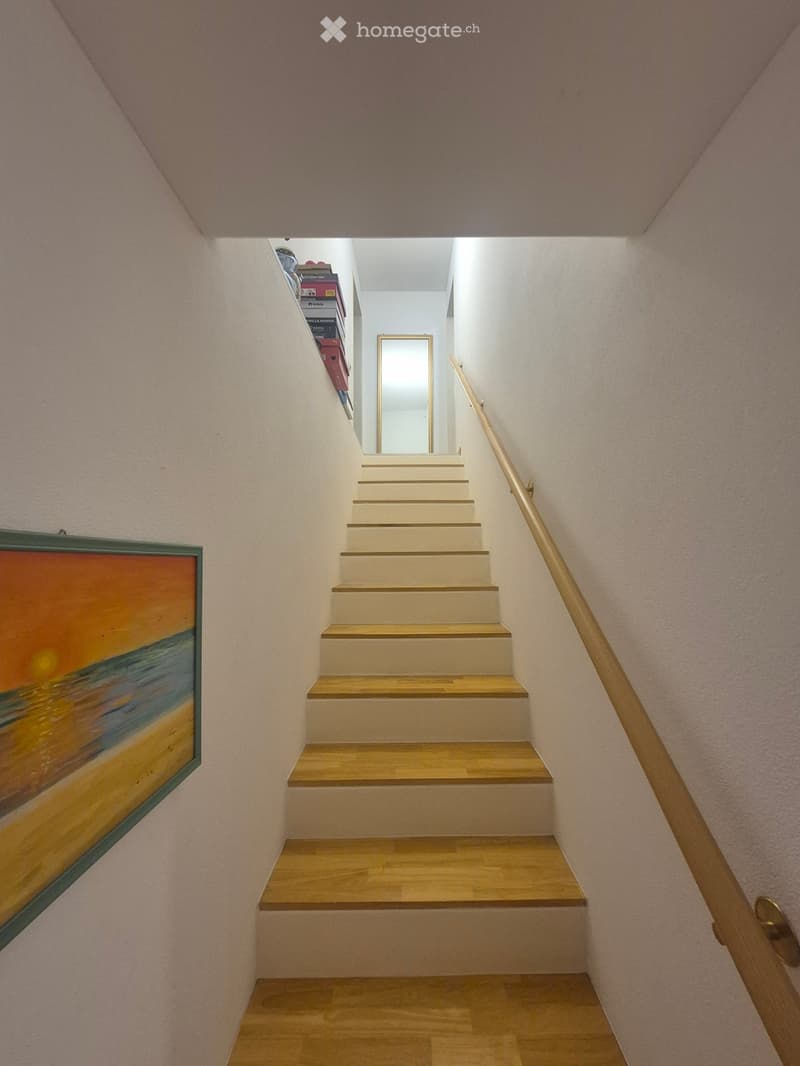 A narrow staircase with wooden steps and a wooden handrail, leading upwards. The walls are painted in a light color, and there is a framed painting on the left wall. A small shelf with books is visible on the left side of the staircase.
