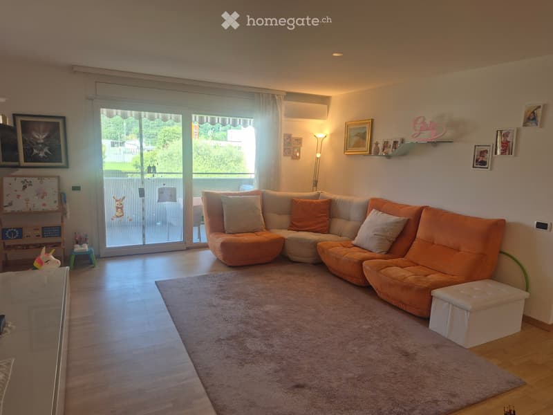 Modern living room with a large orange sectional sofa, a glass coffee table, a floor lamp, and a shelf with picture frames and decorative items. The room features wooden flooring, a large window with a view, and a small child's play area in the corner.