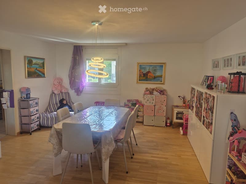 Brightly lit dining room with wooden flooring, a dining table with chairs, a sofa, storage units, toys, and picture frames on the walls.