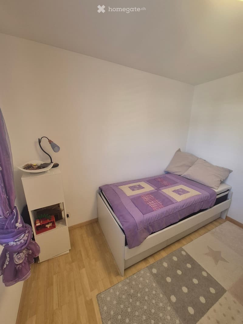 A small bedroom with a single bed, nightstand, lamp, and a purple curtain. The bed has a purple quilt and matching pillows, and there is a small rug on the floor.