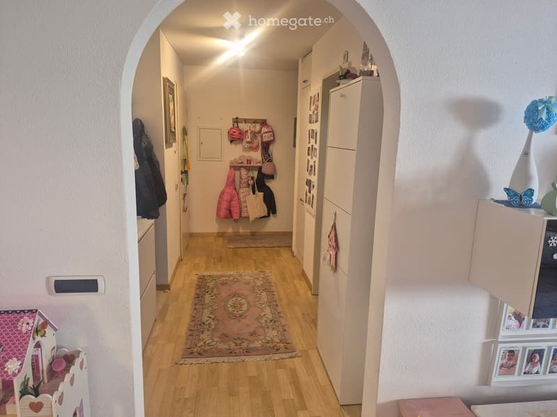 A well-lit hallway with wooden flooring, white walls, and a small patterned rug. Various items are visible, including coats hanging on hooks, a small toy house, framed pictures, and decorative vases.