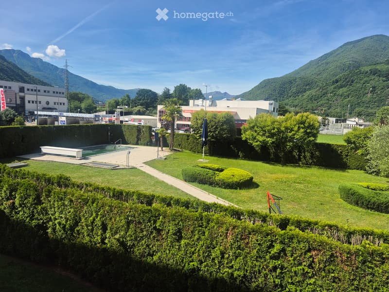 Well-maintained garden with trimmed hedges, a small swimming pool, and a pathway leading to surrounding buildings and mountains.