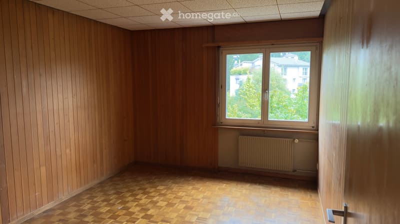 Empty room with wooden panel walls, herringbone wooden flooring, a window with white frame, and a radiator below the window.