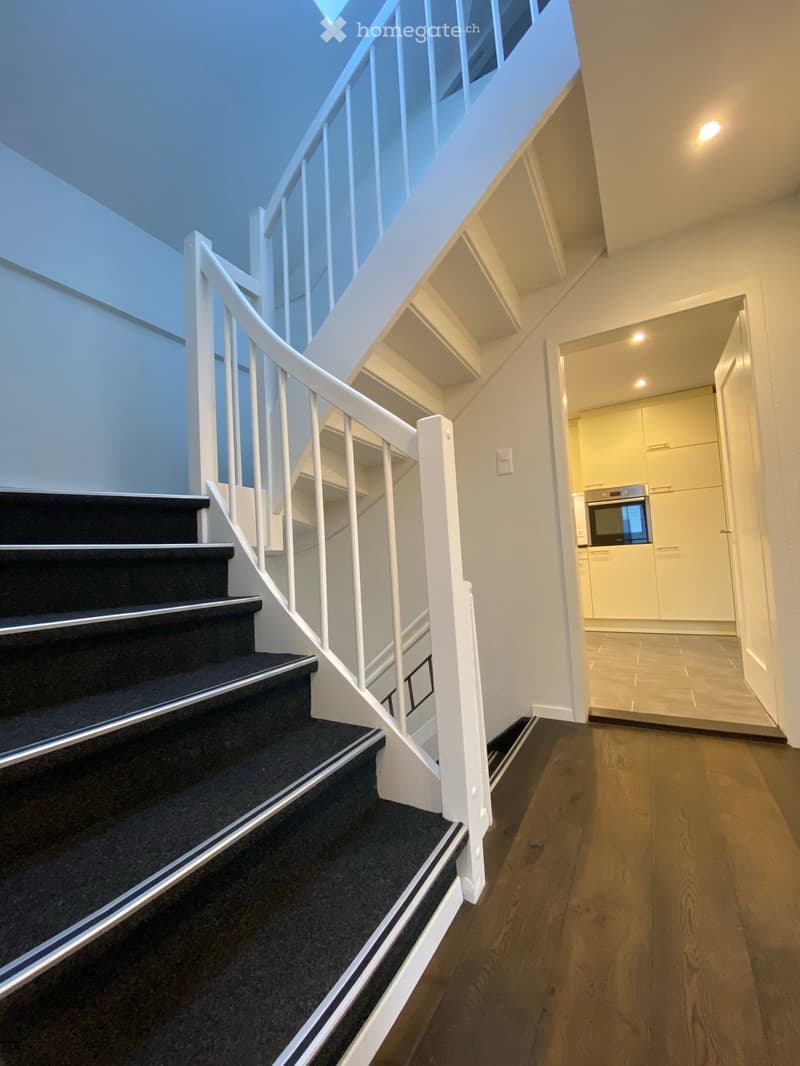 Modern hallway with wooden flooring, white staircase with black carpeted steps, white railings, and a view into a kitchen area with white cabinets and stainless steel appliances.