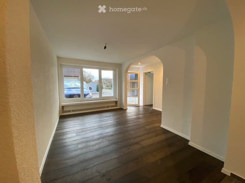 Empty hallway with wooden flooring, white walls, and a large window showing a parked car outside. The hallway has an arched doorway leading to another room.