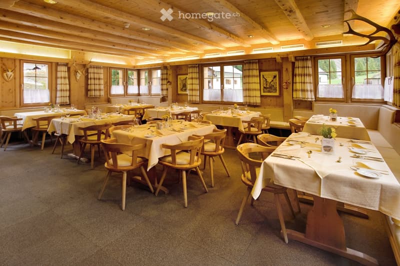 A cozy restaurant with wooden tables and chairs, set with plates, cutlery, and glasses, large windows with curtains, and wooden walls with mounted animal heads and artwork.