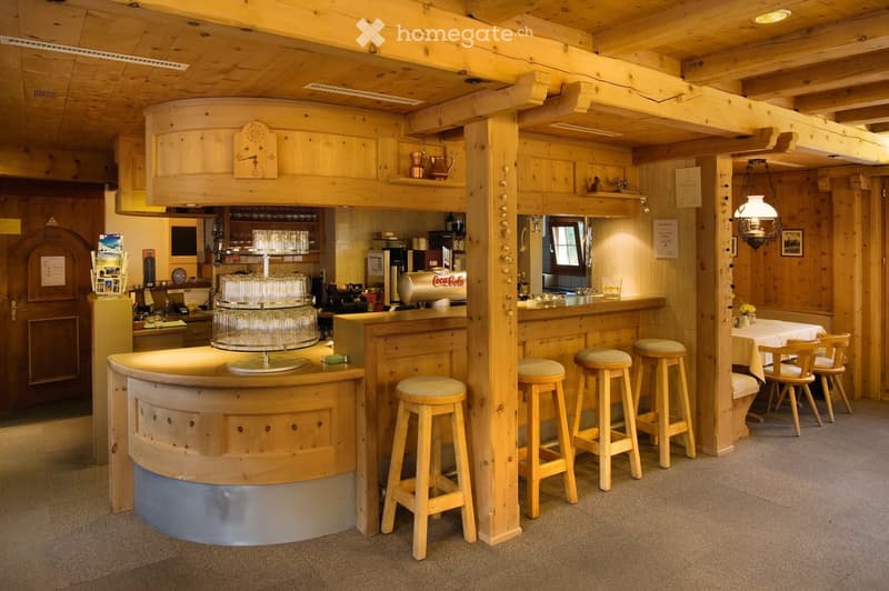 A cozy wooden bar area with a curved wooden counter, several wooden stools, a small table with chairs, and various bar equipment including a refrigerator, screen, and decorative items.