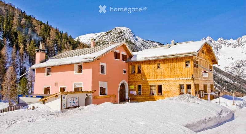 Two-story building with pink and wooden facades, multiple windows, and a snow-covered roof, situated in a mountainous area with snow-covered ground.