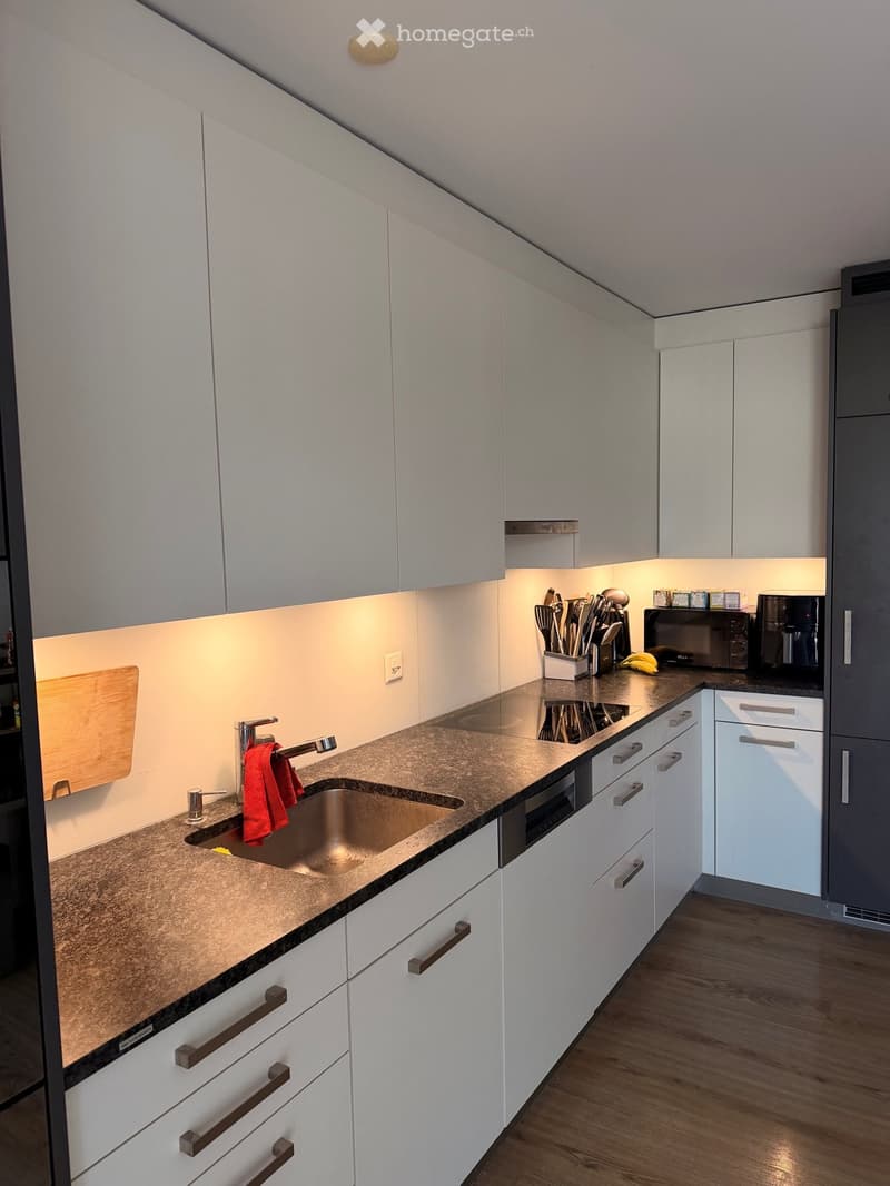 Modern kitchen with white cabinets, granite countertop, stainless steel sink, and various appliances including a microwave and coffee maker. Wooden cutting board and utensils are visible on the counter.