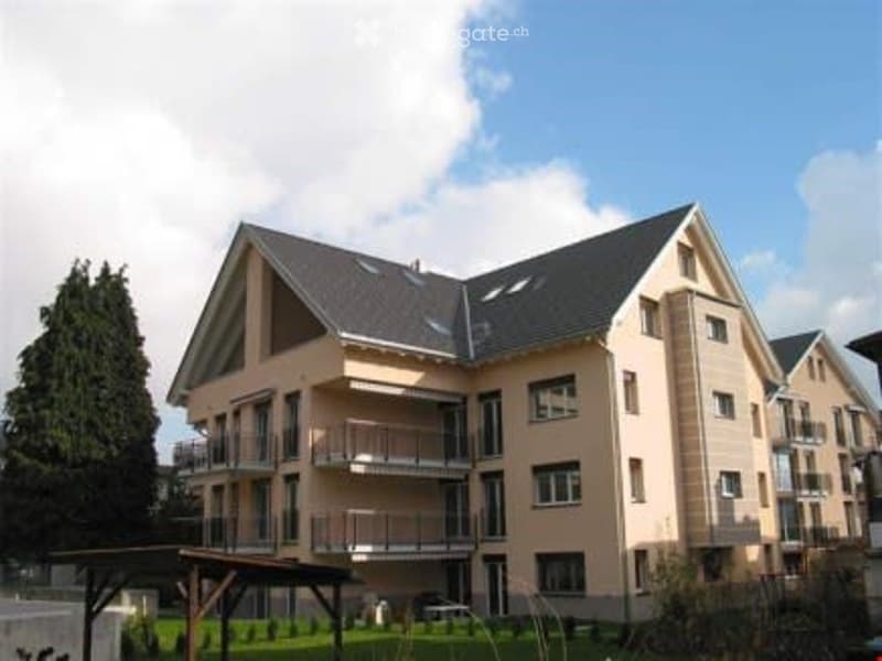Modern multi-story residential building with beige facade, dark gray roof, multiple balconies, and surrounding greenery.