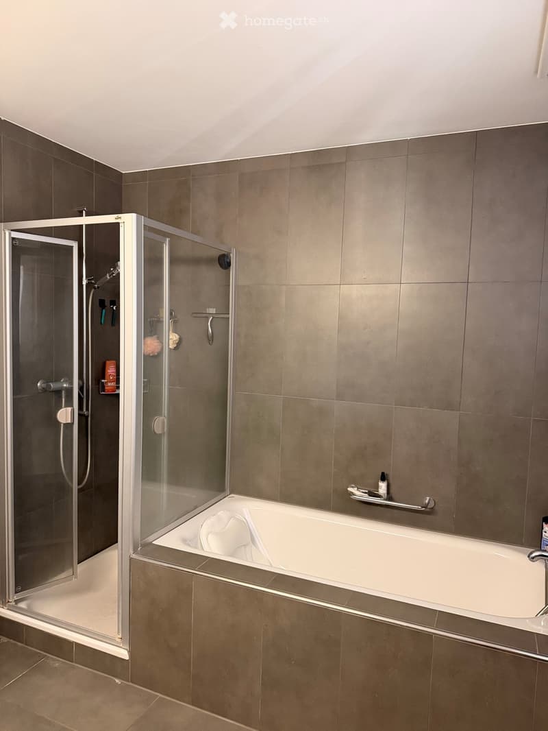 Modern bathroom with a bathtub, glass-enclosed shower, and a handrail beside the tub. The walls are tiled in a neutral gray color, and the floor is tiled in a light brown color.