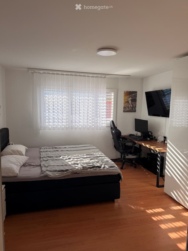 A modern bedroom with a bed, desk, chair, computer, and TV screen. The room features wooden flooring, a large window with sheer curtains, and a painting on the wall.