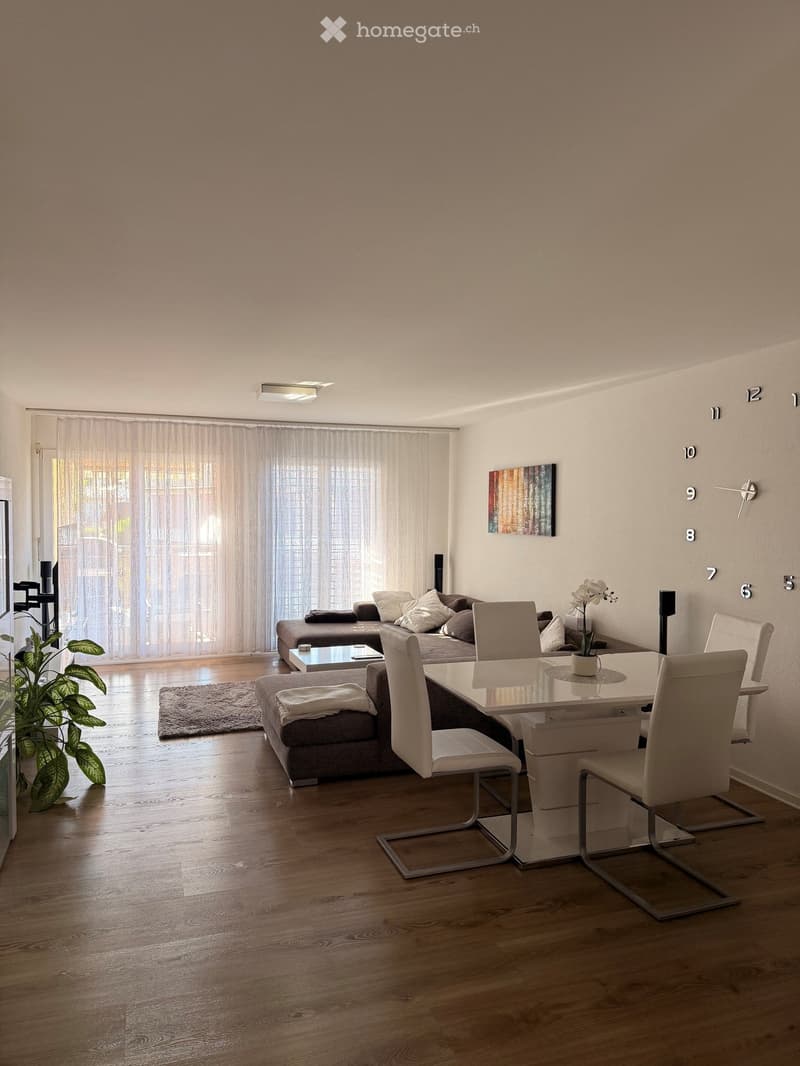 Modern living and dining area with a grey sectional sofa, white dining chairs, a glass dining table, a coffee table, a potted plant, and a wall-mounted clock.