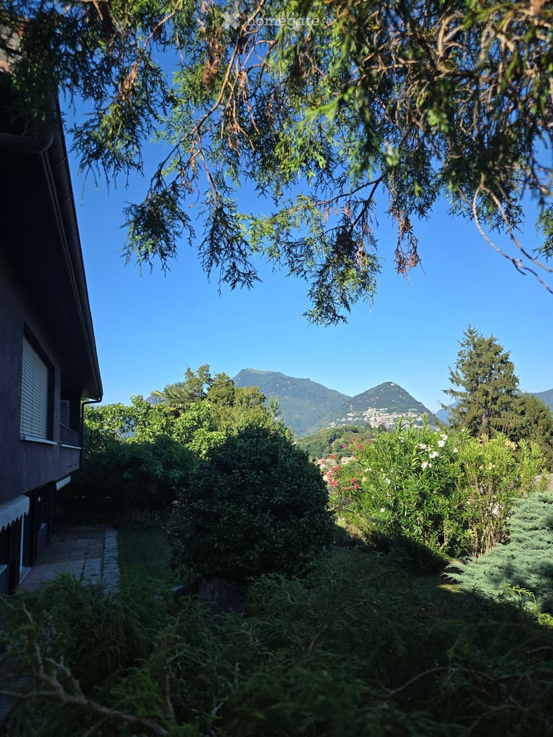 A lush garden area with dense greenery, flowers, and a pathway leading to a house, with mountains visible in the background.