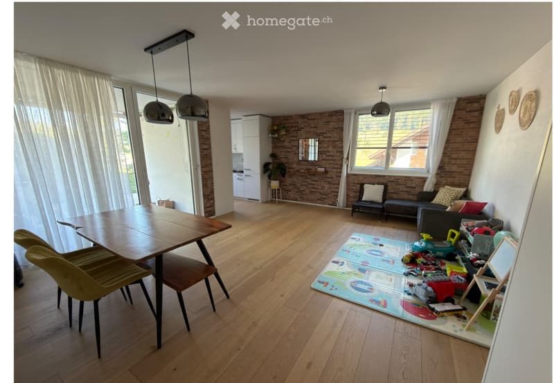 Open-plan living and dining area with wooden flooring, pendant lights, a sofa, a dining table with chairs, a small stool, a mirror, and a play area with toys.