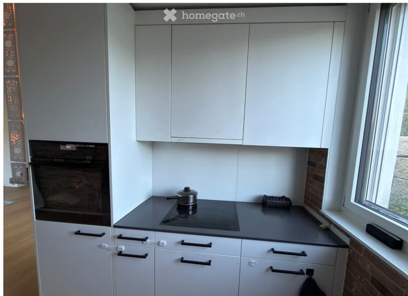 Modern kitchen with white cabinets, black countertop, induction cooktop, and a pot on the stove. The oven is integrated into the cabinetry, and the space features a large window with a view of greenery.