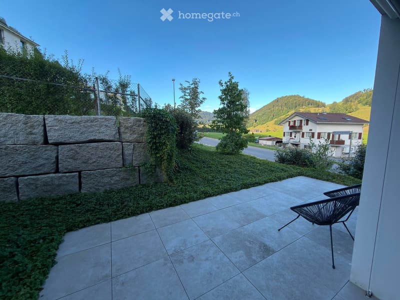 Balcony with tiled floor, stone wall, greenery, and a single black chair.