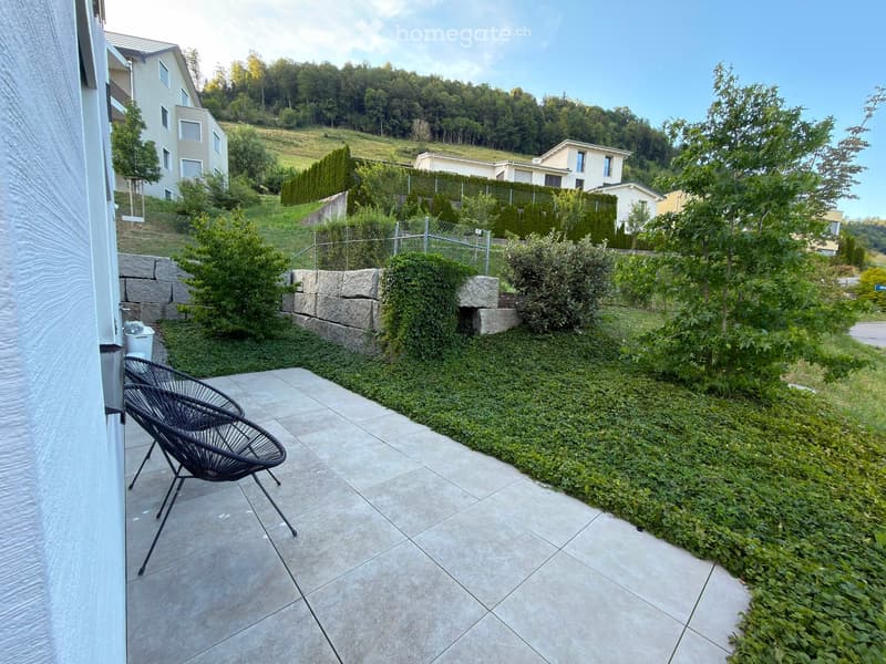 A modern outdoor patio with tiled flooring, two black wicker chairs, and a neatly trimmed hedge border. The area is bordered by a stone wall and overlooks a landscaped garden with trees and neighboring houses in the background.