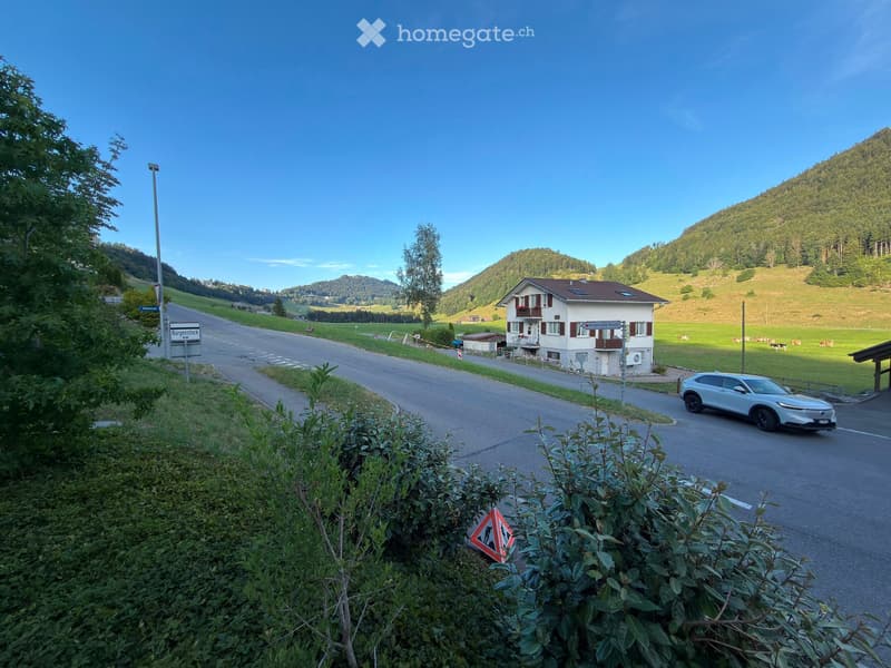 A two-story house with a brown roof and white walls, situated on a road surrounded by green hills and a clear blue sky.