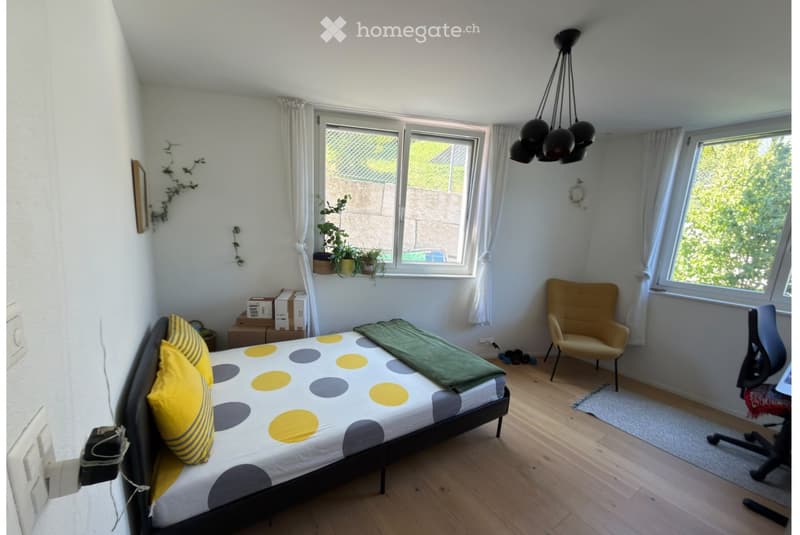 Cozy bedroom with wooden flooring, large windows with white curtains, a bed with yellow and grey bedding, a yellow armchair, a desk chair, and a hanging lamp.