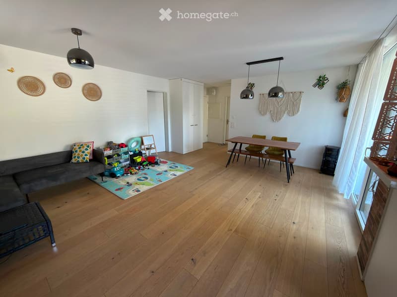 A well-lit living room with wooden flooring, a grey sofa, a dining table with chairs, a cabinet, and various toys on the floor.