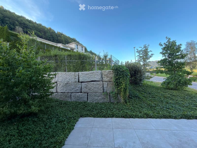 Garden area with a stone retaining wall, ivy-covered fence, tiled pathway, and surrounding greenery.