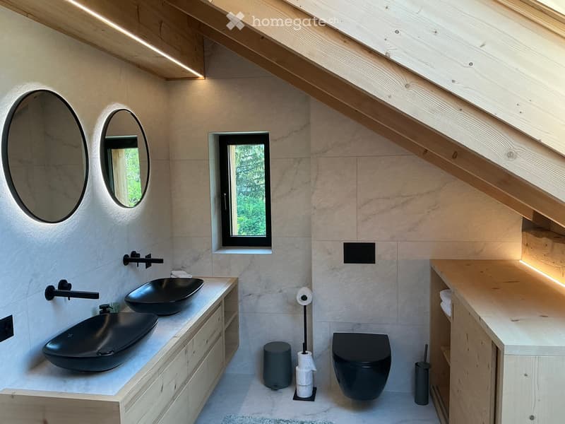 Modern bathroom with sleek black sinks, wooden cabinets, and large mirrors. Features a skylight window and exposed wooden beams on the ceiling.